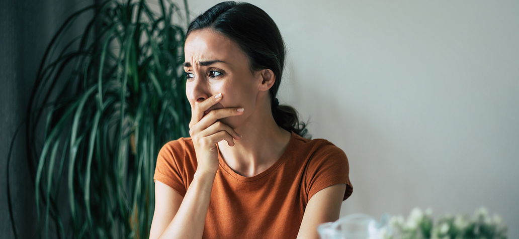 Sad, frustrated young brunette woman is crying with smartphone in hands while she sitting on the chair at apartment